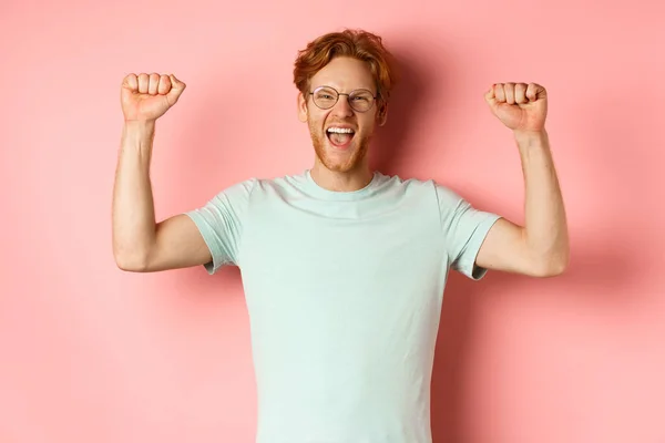 Feliz ganador con el pelo rojo y la barba celebrando la victoria, gritando sí con alegría y levantando las manos, disfrutar del éxito, de pie sobre el fondo rosa — Foto de Stock