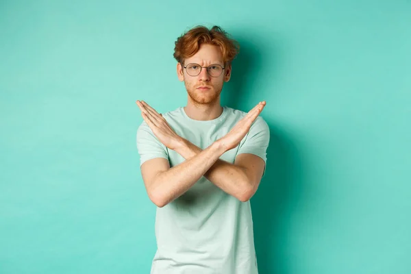 Serious and confident redhead man in t-shirt and glasses saying no, showing cross gesture to stop you, refucing or declining something, standing over turquoise background