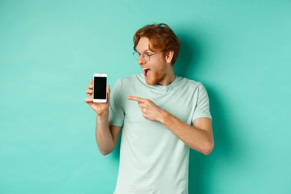 Mira esto. Guapo pelirrojo en gafas apuntando con el dedo a la pantalla del teléfono inteligente en blanco, mostrando promoción en línea, de pie sorprendido sobre fondo turquesa — Foto de Stock