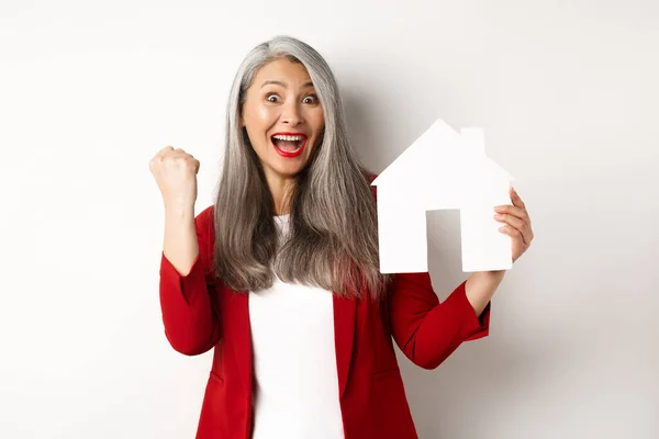 Glückliche asiatische Großmutter zeigt Papierhaus-Ausschnitt und Faust-Pump-Geste, schreit vor Freude ja, kauft Immobilien, steht vor weißem Hintergrund — Stockfoto
