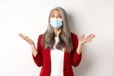 Coronavirus ve iş konsepti. Yüz maskesi takmış Asyalı bir iş kadını ellerini yana açmış ve sol üst köşede şoke olmuş bir şekilde beyaz arka plan izliyor.