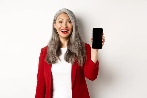 Glada asiatiska kvinnliga entreprenör visar mobiltelefon skärm, leende road, står över vit bakgrund — Stockfoto