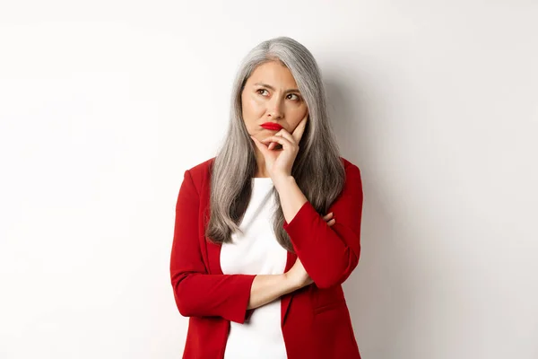 Problémová asijská podnikatelka v červeném saku a rtech, trucuje a dívá se otráveně vlevo, stojí na bílém pozadí — Stock fotografie