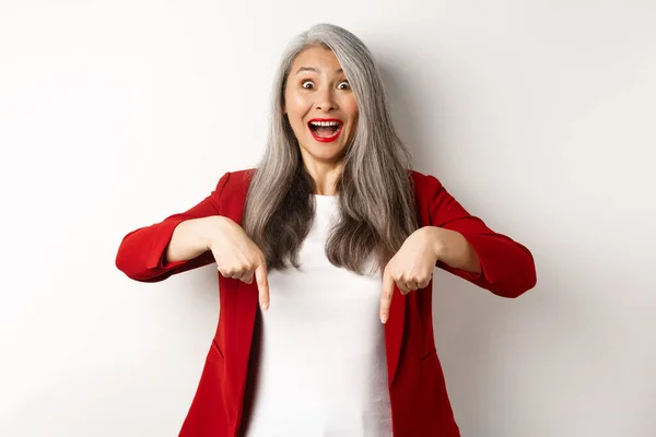 Portrait einer glücklichen asiatischen Dame in rotem Blazer mit Logo, Zeigefinger nach unten und fröhliches Lächeln, überprüfen Sie diese Geste, weißer Hintergrund — Stockfoto