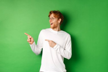Şaşırmış gözlüklü kızıl saçlı adam tanıtımı kontrol ediyor, şaşırmış bir şekilde sola bakıyor, yeşil arka planda uzun kollu tişörtle duruyor.
