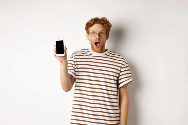 Concepto de tecnología y comercio electrónico. Sorprendido y sorprendido pelirrojo chequeando la promoción en línea, mostrando la pantalla del teléfono inteligente en blanco y la mandíbula caída, fondo blanco — Foto de Stock