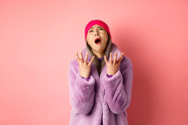 Miserable asiático senior mujer en elegante púrpura abrigo de piel sintética, rogando a Dios y preguntando por qué, estrechando la mano y mirando hacia arriba desesperado, de pie sobre el fondo rosa — Foto de Stock