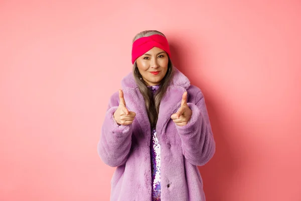 Mode- und Einkaufskonzept. Coole asiatische Seniorin in stylischem Kunstpelzmantel zeigt mit den Fingern in die Kamera und bittet dich, das Promo-Angebot zu prüfen, steht über rosa Hintergrund — Stockfoto