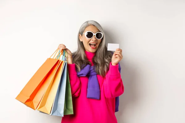 Stilvolle asiatische Großmutter mit Sonnenbrille, die im Urlaub einkaufen geht, mit Papiertüten und Plastikkreditkarte vor weißem Hintergrund — Stockfoto