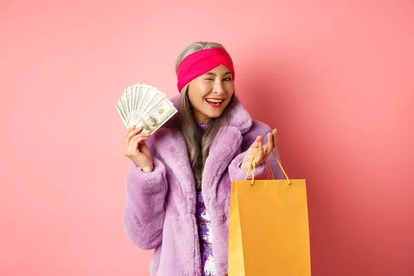 Reiche und modische asiatische Seniorin verschwendet Geld in Geschäften, hält Einkaufstasche und Dollars in der Hand, zwinkert glücklich in die Kamera, rosa Hintergrund — Stockfoto