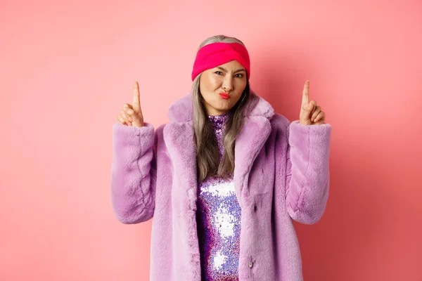 Decepcionado elegante asiática dama señalando los dedos hacia arriba. Mujer mayor en abrigo de piel falsa de moda haciendo muecas escépticas y señalando los dedos hacia arriba, no le gusta la promoción, de pie sobre el fondo rosa —  Fotos de Stock