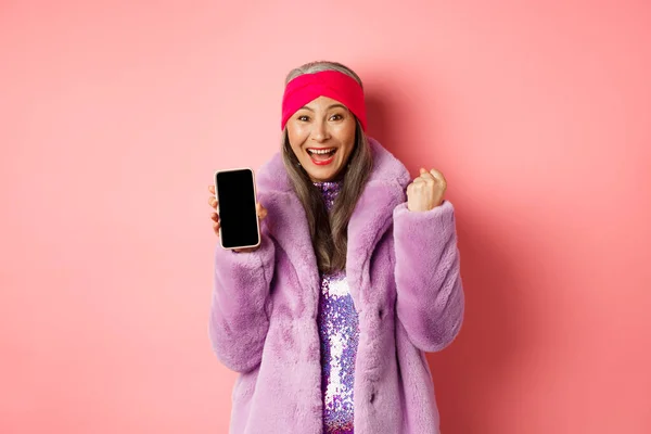 Online-Shopping und Modekonzept. Glückliche asiatische Seniorin gewinnt Preis im Internet, zeigt leeren Handy-Bildschirm und macht Faustpumpe, schreit vor Freude, steht über rosa Hintergrund — Stockfoto