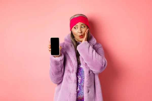 Valentinstag Shopping-Konzept. Schöne asiatische Seniorin zeigt Smartphone-Bildschirm und schaut aufgeregt auf Display, schaut sich Online-Angebot an, pinkfarbener Hintergrund — Stockfoto