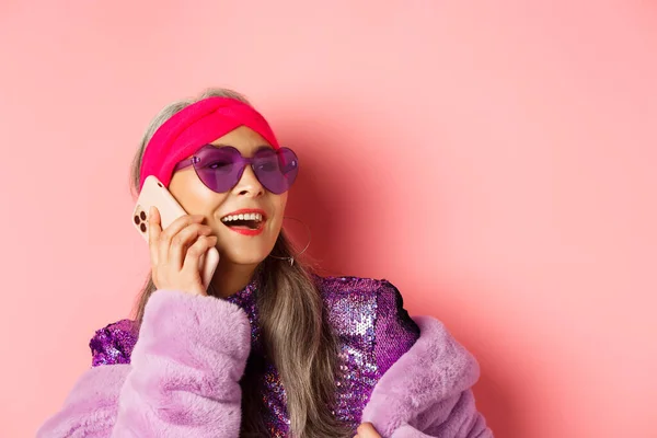 Мода. Крупный план модной азиатской пожилой женщины в солнцезащитных очках и диско-костюме, разговаривающей по телефону, смеющейся и улыбающейся, стоящей на розовом фоне — стоковое фото
