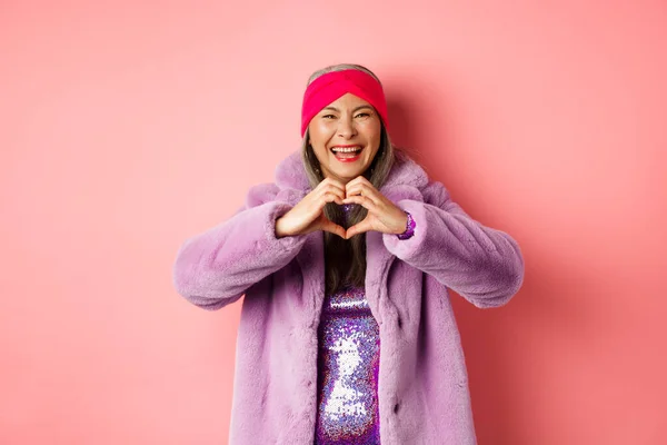 Conceito de amor e moda. feliz ásia sênior fêmea mostrando coração sinal e sorrindo para câmara eu amo você gesto de pé contra rosa fundo — Fotografia de Stock