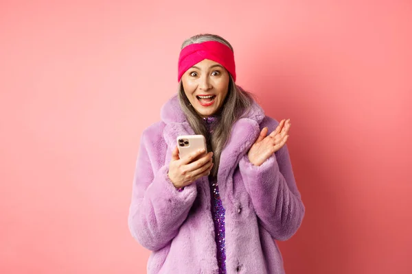 Online-Shopping und Modekonzept. Asiatische reife Frau sieht aufgeregt aus, als sie Internet-Promo-Angebot auf dem Smartphone anschaut, glücklich in die Kamera schaut, pinkfarbener Hintergrund — Stockfoto