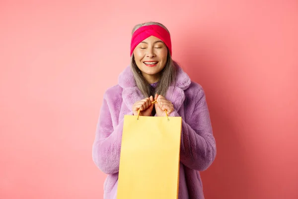 Shopping- und Modekonzept. Glückliche asiatische Seniorin in stilvoller Kleidung sieht glücklich und erfreut aus, steht mit gelber Papiertüte und lächelndem, rosa Hintergrund — Stockfoto