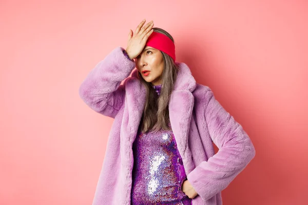 Conceito de moda e compras. Mulher sênior irritado rolar os olhos e fazer facepalm de algo coxo, de pé em casaco de inverno roxo na moda e vestido brilhante, fundo rosa — Fotografia de Stock