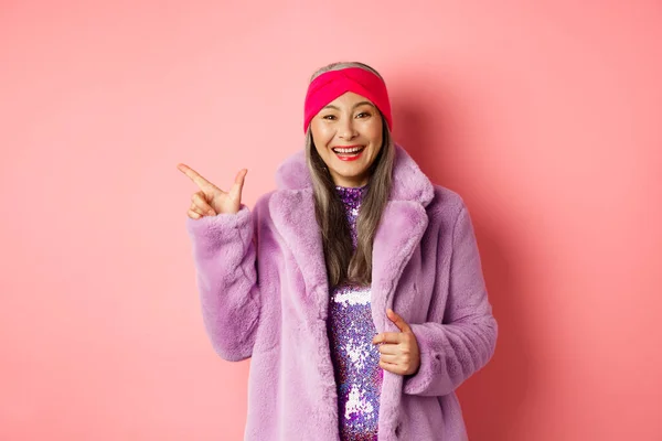 Mode- und Einkaufskonzept. Glückliche asiatische Seniorin checkt trendige Promotion, zeigt mit dem Finger nach links auf das Logo und lächelt, trägt winterlichen Kunstpelzmantel und Stirnband — Stockfoto