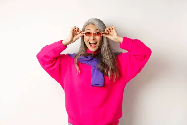 Modische asiatische Seniorin probiert neue Sonnenbrille aus, schaut erstaunt und fröhlich in die Kamera, steht im trendigen Pullover vor weißem Hintergrund — Stockfoto