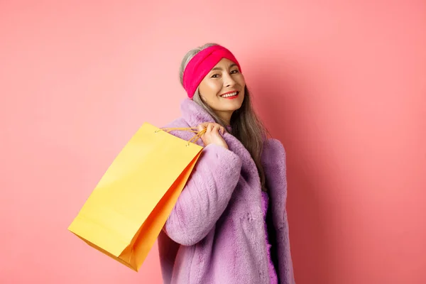 Stilvolle asiatische Seniorin geht einkaufen, trägt Papiertüte auf der Schulter und lächelt glücklich in die Kamera, steht über rosa Hintergrund — Stockfoto