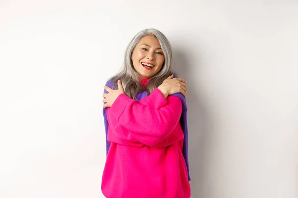 Concepto de San Valentín y vacaciones. Preciosa asiática senior mujer en rosa suéter abrazándose con los ojos cerrados, sonriendo, de pie sobre fondo blanco — Foto de Stock