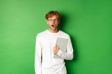 Şaşırmış genç bir adam dizüstü bilgisayar tutuyor ve kameraya bakıyor, gözlüklü ve beyaz tişörtlü, yeşil arka planda duruyor.