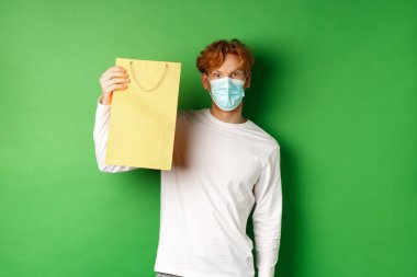 Covid-19 ve pandemik konsept. Yüzü maskeli, alışveriş torbasını gösteren ve yeşil arka planda duran kameraya bakan şaşırmış bir adam.