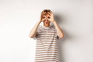 Etkilenmiş kızıl saçlı adam reklam teklifini kontrol ediyor, kameraya el dürbününden bakıyor, şaşkın bir şekilde gülümsüyor, beyaz arka plan.