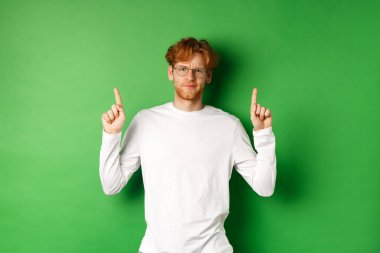 Kızıl saçlı ve gözlüklü hoşnutsuz genç adam, parmaklarını kaldırıyor ve kaşlarını çatıyor, hayal kırıklığına uğruyor, yeşil arka planda duruyor.