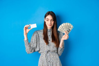 Alışverişe. Elbiseli güzel kız yana bakıyor ve gülümsüyor dalgın dalgın, para ve kredi kartı gösteriyor, mavi arka planda duruyor.