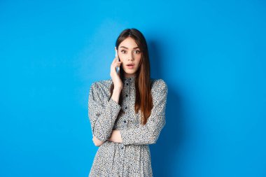 Şok olmuş bir kız cep telefonuyla konuşuyor, kameraya hayret ediyor, biriyle konuşuyor ve şok edici haberler duyuyor, mavi arka planda giyinmiş duruyor.