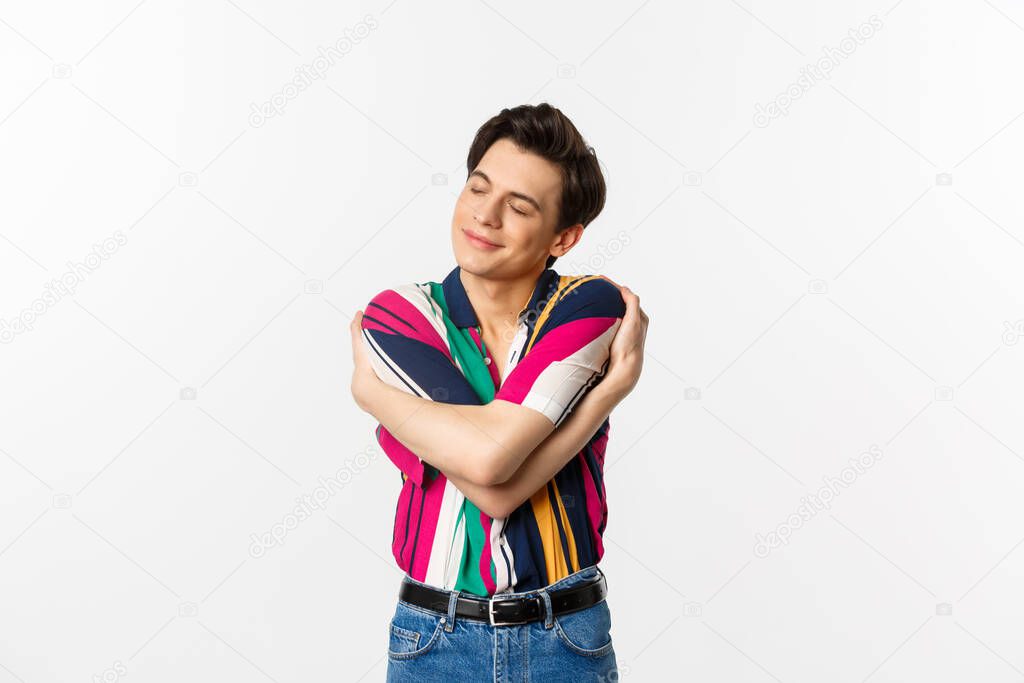 Happy young man hugging himself with eyes closed, smiling pleased, dreaming about something, standing over white background