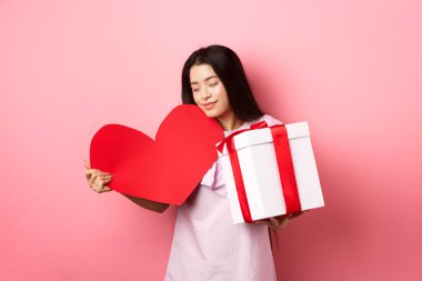 Sevgililer Günü konsepti. Romantik Asyalı genç kız beyaz bir günde sevgilisinden hediyeler alır. Büyük kırmızı kalp kartına sarılır ve kutuda sunulur. Güler yüzlü, pembe arka plan.