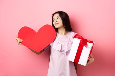 Sevgililer Günü konsepti. Aşk hediyeleri tutan romantik kız, büyük kırmızı kalp kartı ve hediye kutusu, rüya gibi bakışlar, aşık olmak, şefkatle gülümsemek, pembe arka plan