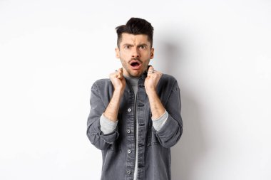 Şok olmuş genç adam şaşkın ve dargın görünüyor, kameraya bakıp somurtuyor, kötü haberleri duyuyor, beyaz arka planda duruyor.