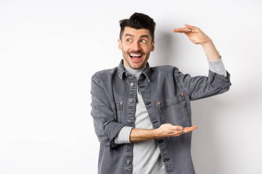 Heyecanlı adam logoya bakıyor ve büyük bir şey gösteriyor, büyük bir nesne şekillendiriyor ve şaşkın bir şekilde gülümsüyor, beyaz arka planda kot ceketle duruyor.