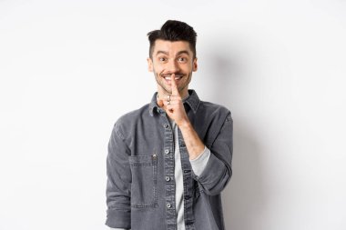 Mutlu adam kameraya şşşş yapıyor, parmakları dudaklarında gülümsüyor, sessiz kalmak istiyor, büyük bir sır veriyor ya da sürpriz yapıyor, beyaz arka planda duruyor.
