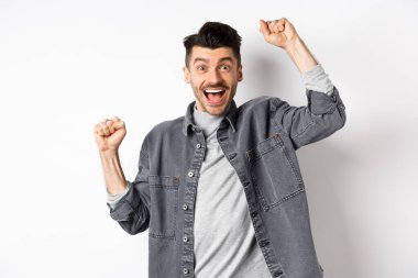 Heyecanlı adam sevinçten bağırıyor, ödül kazanıyor ve seviniyor, ellerini kaldırıyor ve evet diye bağırıyor tatmin olmuş yüzle, zaferle, hedefe ulaşıyor, beyaz arka planda duruyor.