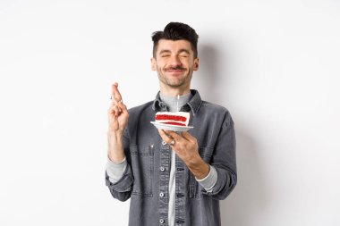 Umut dolu doğum günü çocuğu dua ediyor, partide doğum günü pastası tutuyor, beyaz arka planda duruyor.