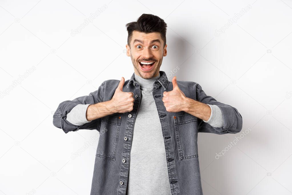 Sounds great. Excited young man showing thumbs up and smiling happy, recommending cool thing, praise nice job, standing on white background