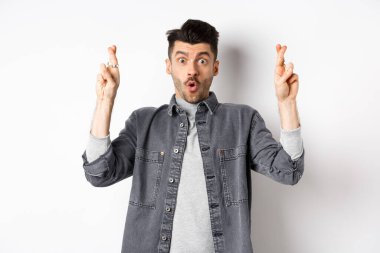 Heyecanlı genç adam şans dilemek ve nefes nefese kalmak için dua ediyor. Önemli haberleri bekliyor, yalvarıyor ya da dua ediyor, beyaz arka planda duruyor.