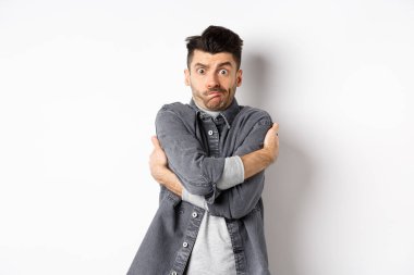 Komik genç adam kendini kucaklıyor, yüzünü buruşturuyor, utanmış ve teşhir edilmiş hissediyor, vücudunu insanlardan gizliyor, beyaz arka plan.