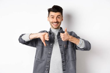 Mutlu gülümseyen adam ortalama bir hareket gösteriyor, baş parmağını kaldırıyor, karar veremiyor, bir şeyi orta derecede yargılıyor, beyaz arka planda duruyor.