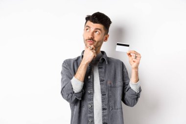 Sol üst köşedeki logoya bakıp elinde plastik kredi kartı tutan, alışveriş yapmayı düşünen, beyaz arka planda duran düşünceli bir adam.