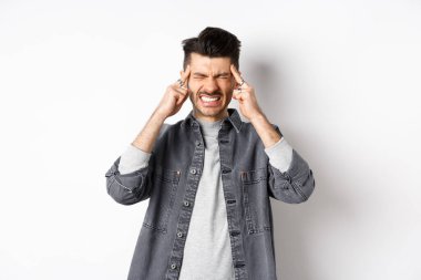 Baş ağrısı çeken, baş ağrısından dişlerini sıkan, baş şakaklarına dokunan, migren ağrısı çeken, beyaz arka planda duran, ağrı kesicilere ihtiyacı olan bir adam.