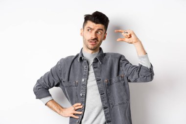 Düşünceli genç adam yana bakıyor ve düşünüyor, küçük bir nesne gösteriyor, küçük ya da küçük bir şeyi düşünüyor, beyaz arka planda duruyor.