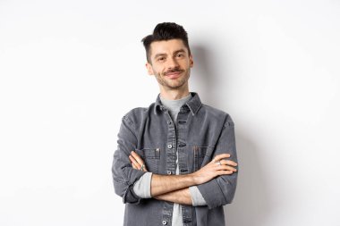 Gururlu yakışıklı adam tatmin olmuş görünüyor, göğsünde çapraz kollar ve kameraya gülümsüyor, iyi bir şeye bakıyor, beyaz arka planda duruyor.