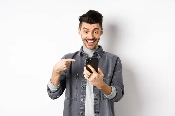 Spännande stilig man som visar coola nyheter på telefon, pekar på smartphone och ler glad, står mot vit bakgrund — Stockfoto