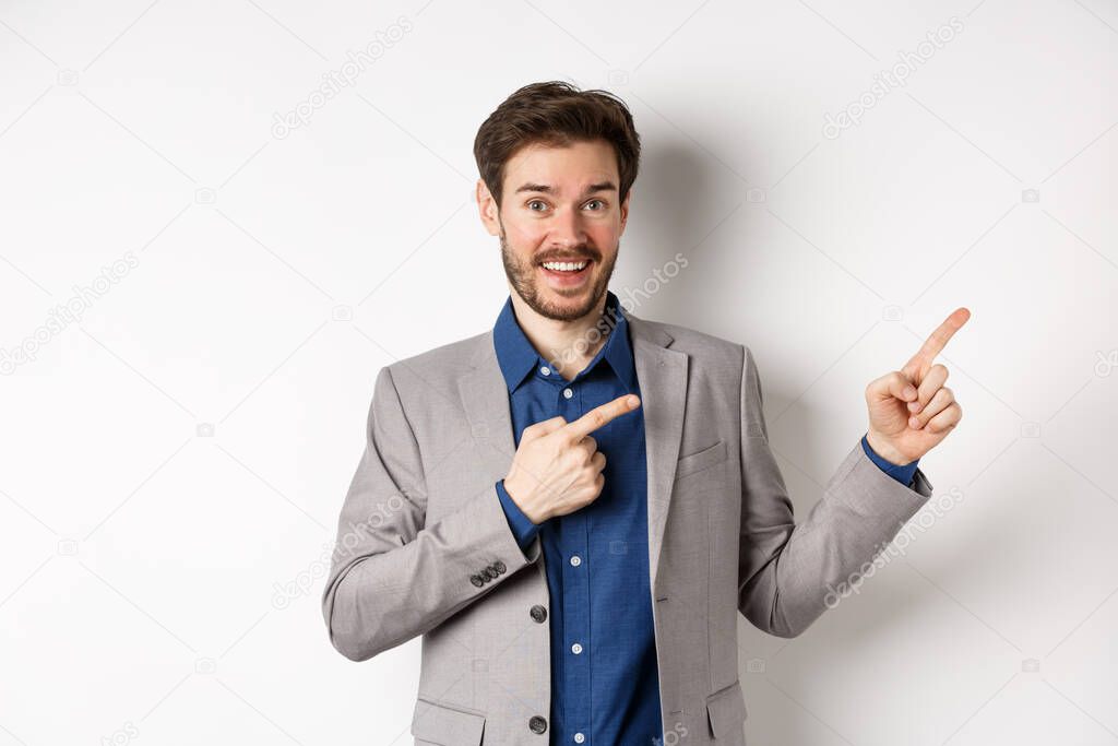 Happy businessman in suit pointing fingers right and smiling excited, showing special deal, standing on white background